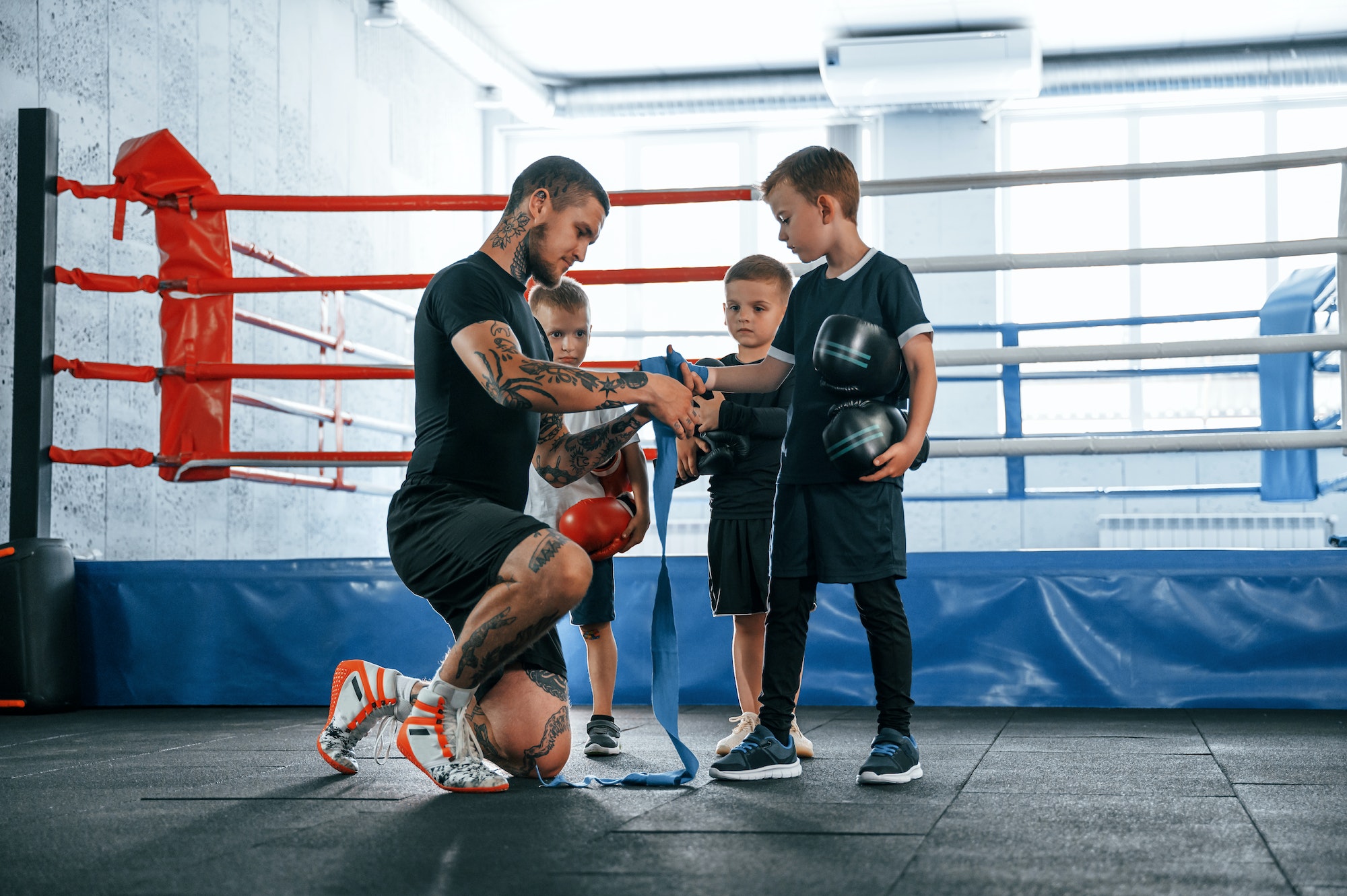 Wearing blue hand wraps. Young tattooed coach teaching the kids boxing techniques